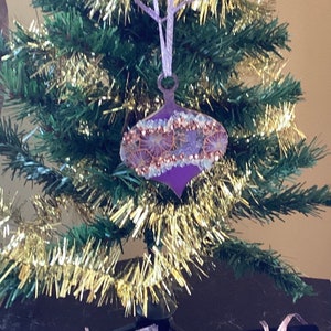 May include: Four purple Christmas ornaments with gold and white accents. The ornaments are decorated with glitter and have a teardrop shape.