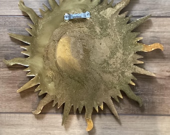 May include: A gold-colored sun-shaped wall hanging with a textured, glittery finish and radiating pointed rays. The back of the object features a central circular indentation and a silver-toned metal sawtooth hanger attached for mounting purposes.