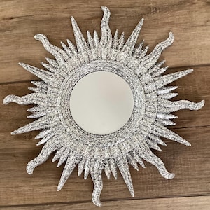 May include: A silver glitter sunburst mirror with a round mirror in the centre. The mirror has a textured, raised design.