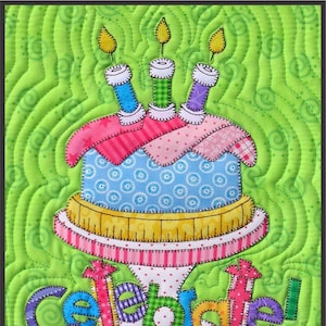 May include: A colourful quilted birthday cake with three lit candles and the word "Celebrate!" in a whimsical font. The cake is on a pink and white striped stand with polka dots.