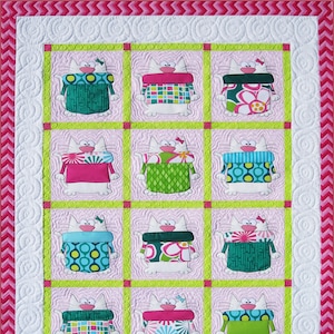 Puede incluir: Una colcha con fondo rosa y borde blanco. La colcha presenta una cuadrícula de nueve cuadrados, cada uno con un gato blanco que lleva un suéter con diferentes patrones. Los suéteres son de varios colores, incluyendo verde, azul, rosa y blanco.