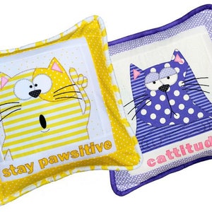 May include: Two decorative throw pillows with embroidered cat faces and text. The yellow pillow has a yellow and white striped cat with the text "stay pawsitive". The purple pillow has a purple and white polka dot cat with the text "cattitude".