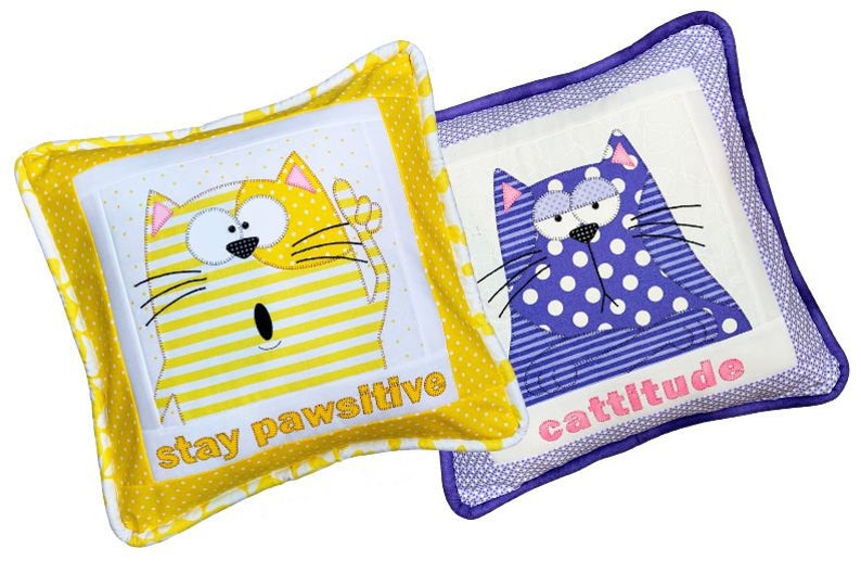 May include: Two decorative throw pillows with embroidered cat faces and text. The yellow pillow has a yellow and white striped cat with the text "stay pawsitive". The purple pillow has a purple and white polka dot cat with the text "cattitude".