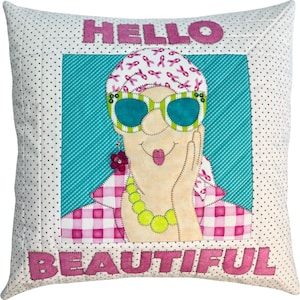 Puede incluir: Una almohada acolchada rosa y blanca con las palabras "HELLO BEAUTIFUL" en letras rosas. El centro de la almohada presenta una imagen de dibujos animados de una mujer que lleva ropa a cuadros rosa y blanca, gafas de sol y una diadema rosa con cintas rosas.