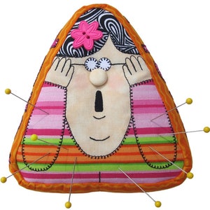 May include: A triangular pincushion shaped like a person with pink and green striped clothing, a pink flower, and white eyes. The pincushion has yellow pins sticking out of it.