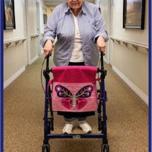 Op de afbeelding: Een blauwe rollator met een felroze stoffen tas aan de voorzijde. De tas is voorzien van een grote, kleurrijke vlinderapplicatie met roze, paarse en groene details. De tekst "Amy's Mom - Her Design Inspiration." staat onderaan de afbeelding.
