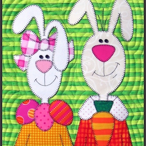 May include: Two white bunny rabbits with embroidered details, one wearing a pink and white gingham bow and the other a green bow tie. The bunny on the left is holding a carrot. The bunnies are on a green and white striped background.