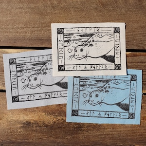 May include: Three fabric patches featuring a black and white illustration of a seal swimming. The patches are in different colours: white, light blue and grey. The text on the patches reads "Sætyrık" and "Fætyr", which are runes.