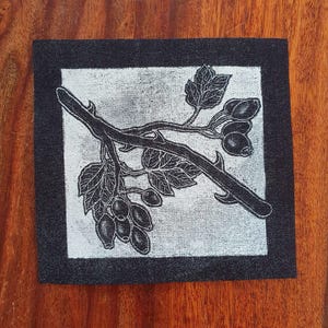 May include: Square artwork with a black and white botanical illustration of a rose hip branch with leaves and berries. The image has a silver background and a black border. The artwork is displayed on a wooden surface.