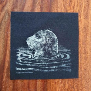 May include: A square, black fabric patch with a white illustration of a seal's head breaking the water's surface. The seal is shown in profile, with detailed shading and whiskers. The water is represented by wavy lines.