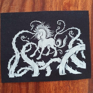 May include: A black rectangular patch featuring a white unicorn design. The unicorn is surrounded by white thorny vines and branches. The patch is likely made of fabric and is set against a wooden background.