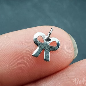 Puede incluir: Un pequeño dije de lazo plateado con un lazo en la parte superior para sujetar a la joyería. El dije se sostiene entre dos dedos sobre un fondo oscuro. El dije mide aproximadamente 1,3 cm de ancho.