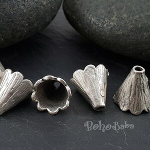 May include: Six silver metal cone-shaped beads with scalloped edges. The beads are arranged in a semi-circle on a dark gray surface.