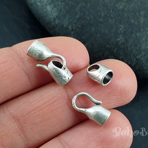 May include: Silver tone metal end caps for jewelry making, shaped like small watering cans with a hook. The end caps are shown on a hand against a dark gray background.