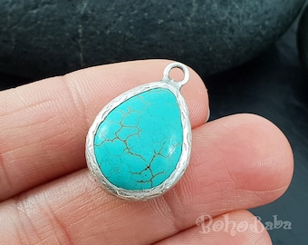 Op de afbeelding: Een druppelvormige hanger met een levendige turkooizen steen gezet in een getextureerde zilverkleurige metalen lijst. De turquoise heeft een natuurlijk, gebarsten patroon. Er zit een kleine lus aan de bovenkant om op te hangen.