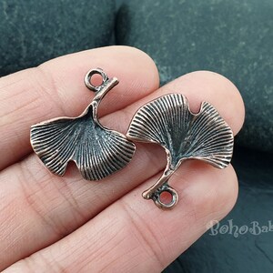 May include: Two copper-colored ginkgo leaf charms. Each charm features a detailed leaf design with a small loop for attaching to jewelry. The charms have a vintage, antique appearance.