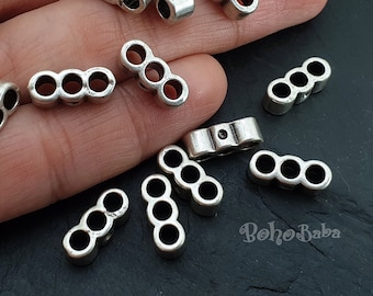 May include: Silver metal spacer beads with three holes each. The beads are arranged on a hand.
