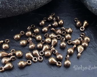 May include: A scattering of antique brass teardrop charms with a loop at the top. The charms are scattered on a dark gray surface.