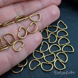 May include: A close-up of a hand holding a pile of gold-colored metal D-rings. The D-rings are all the same size and are arranged in a random pattern. There are about 30 D-rings in the pile.