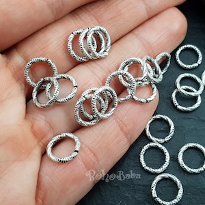 May include: A close-up of a hand holding a pile of silver jump rings. The rings are textured and have a slightly hammered finish.