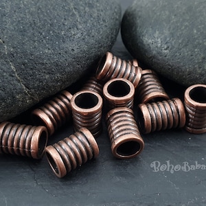 May include: A close-up of a group of 12 copper-coloured metal beads. The beads are cylindrical and have a ridged texture. The beads are arranged in a random pattern on a dark grey surface.