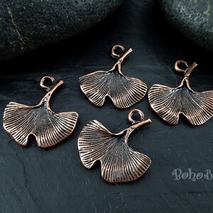 May include: Four copper-colored Ginkgo leaf charms with a textured, veined design. Each charm has a loop at the top for attaching to jewelry. The charms are displayed on a dark, textured surface.