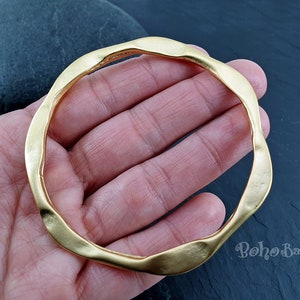 Puede incluir: Un brazalete abierto grueso de color dorado, con una forma ondulada irregular. El brazalete está hecho de metal y tiene un acabado liso y pulido.