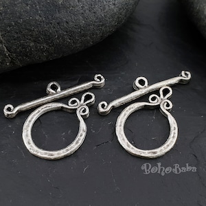 Op de afbeelding: Twee zilverkleurige metalen sluitingen met een gestructureerd oppervlak. Elke sluiting heeft een ronde ring en een rechte stang met decoratieve krulletjes.