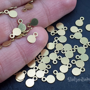 Könnte beinhalten: Nahaufnahme einer Hand, die einen Haufen goldfarbener Metall-Anhänger hält. Die Anhänger sind rund mit einer kleinen Schlaufe oben. Die Anhänger sind auf einer dunklen Oberfläche verstreut.