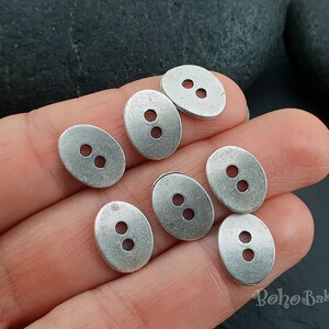 May include: Seven silver oval-shaped buttons with two holes each. The buttons have a slightly textured surface.