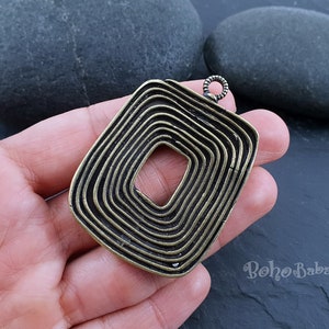 May include: A gold-toned metal pendant with a square shape and a concentric circle design. The pendant has a loop at the top for attaching to a chain or cord.