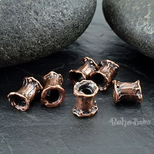 May include: A collection of copper-toned ear gauges with a textured, organic design. The cylindrical plugs are displayed on a dark surface, with two large, dark grey stones in the background. The text "Boho Baba" is visible.