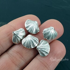 Op de afbeelding: Zes zilverkleurige metalen schelpvormige kralen. De kralen zijn ongeveer 2,5 cm in diameter en hebben een getextureerd oppervlak.