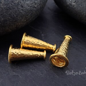 May include: Three gold-toned metal cone-shaped beads with a hammered texture. The beads are lying on a dark grey surface.