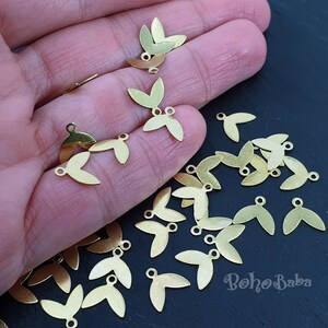 Puede incluir: Primer plano de una mano sosteniendo un montón de abalorios de hoja de metal dorado. Los abalorios tienen forma de hoja con un pequeño bucle en la parte superior para sujetarlos a joyas u otras manualidades.