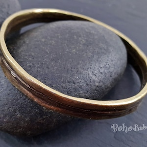 May include: A simple, wide, gold-toned bangle bracelet with a textured, hammered finish. The bracelet is resting on a dark gray stone.