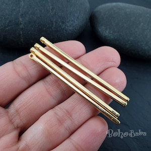 Peut inclure: Trois pendentifs en forme de barre en métal doré, chacun présentant une forme cylindrique lisse et allongée avec une petite boucle circulaire à l'extrémité supérieure pour la fixation. Ces composants sont disposés parallèlement.
