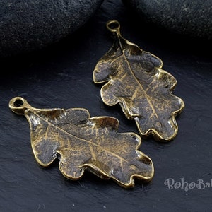 May include: Two antique gold-toned metal oak leaf charms with a loop at the top for hanging. The leaves have a textured, aged finish.