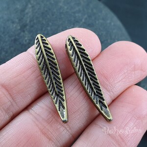 May include: Two antique gold-toned metal feather charms with a detailed etched design.