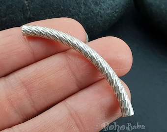 May include: A curved, cylindrical silver-toned metal tube bead featuring a textured, faceted surface pattern. The bead has a hollow center and a polished finish, designed for jewelry making applications. The text BohoBaba is visible in the corner.
