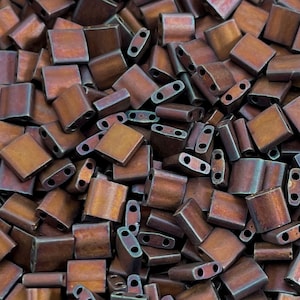 May include: A close-up image of a pile of small, square, copper-colored beads with a metallic sheen. The beads have two holes in each. The beads are arranged in a random pattern.
