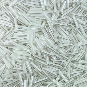 Może przedstawiać: Duża sterta białych szklanych koralików. Koraliki są długie i cienkie, z lekko opalizującym wykończeniem.