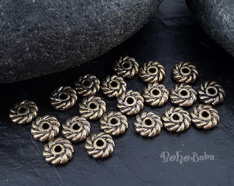 Può includere: Una collezione di 20 perline distanziatrici in metallo color oro antico con un design a corda intrecciata. Le perline sono disposte a semicerchio su una superficie grigio scuro.