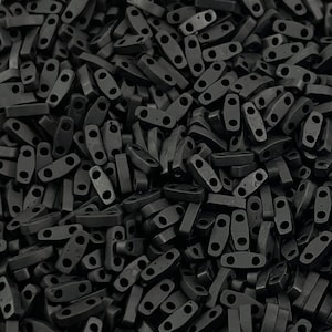 Peut inclure: Un grand tas de perles rectangulaires noires avec deux trous chacune. Les perles sont en matériau mat et ont une texture légèrement rugueuse.