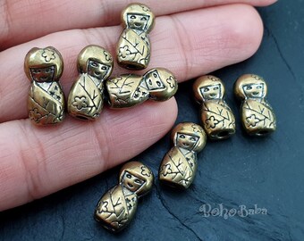 Può includere: Una collezione di otto ciondoli in metallo dorato con design di bambola giapponese. Ogni ciondolo presenta un viso dettagliato e motivi floreali.