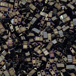 Puede incluir: Una imagen de primer plano de una gran cantidad de pequeñas cuentas de vidrio negras iridiscentes. Las cuentas son de forma rectangular con dos agujeros en cada lado. Las cuentas tienen un brillo metálico y están dispuestas en un patrón aleatorio.