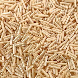 May include: A close-up image of a large quantity of small, light brown glass beads. The beads are cylindrical in shape and have a smooth, glossy finish.