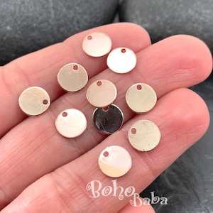 May include: A selection of small, gold-coloured, round metal charms, each with a single hole at the top. The charms are held in a hand, with the text "Boho Baba" visible in the lower right corner.