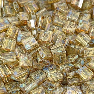 May include: A close-up of a pile of small, square, gold-colored glass beads. The beads have a slightly iridescent finish.