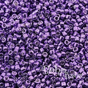 May include: A close-up shot of a pile of small, cylindrical purple beads. The beads are densely packed together, creating a textured surface. The beads have a metallic sheen, reflecting light. The overall effect is a vibrant, shimmering display of color.